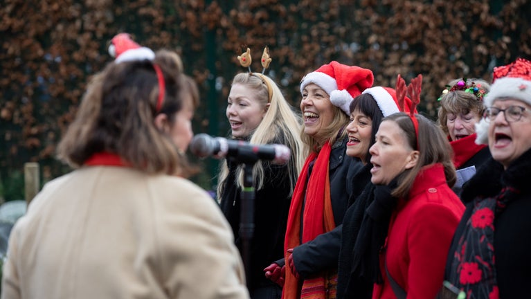 A group of people wearing Christmas hats and antler headbands singing outside.